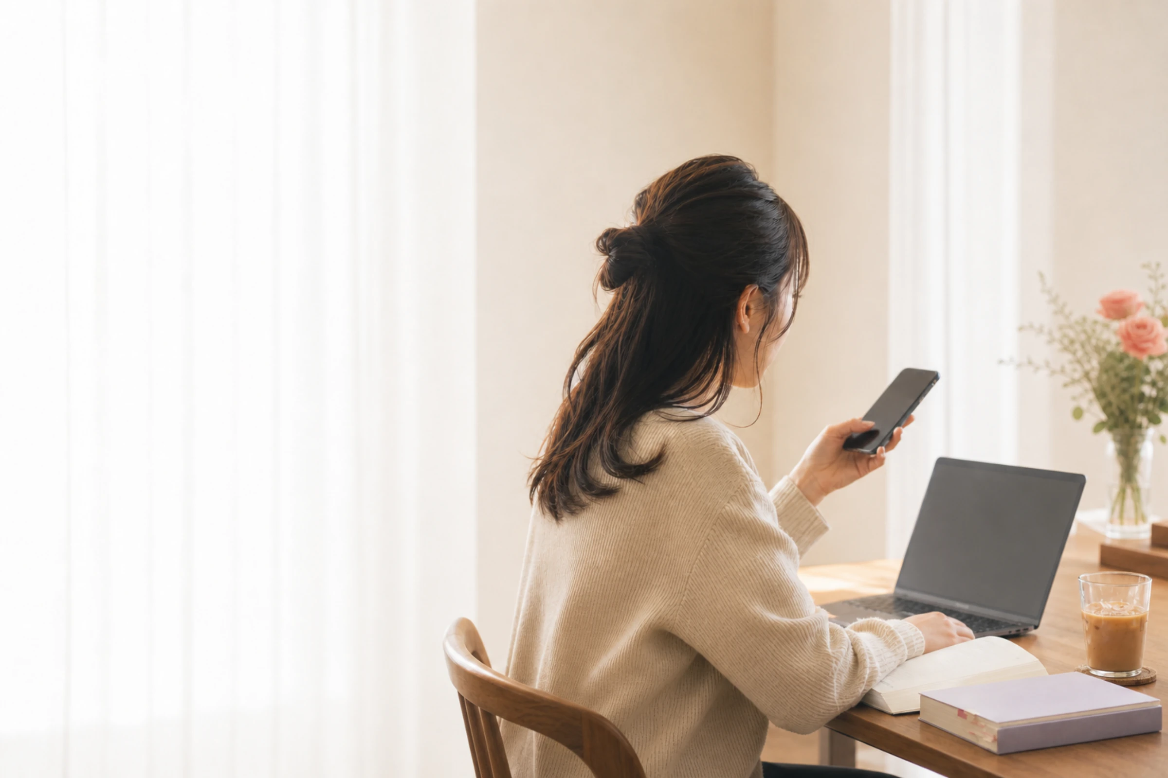 午後の机で、ノートパソコンを開いたままスマホを見るひとり